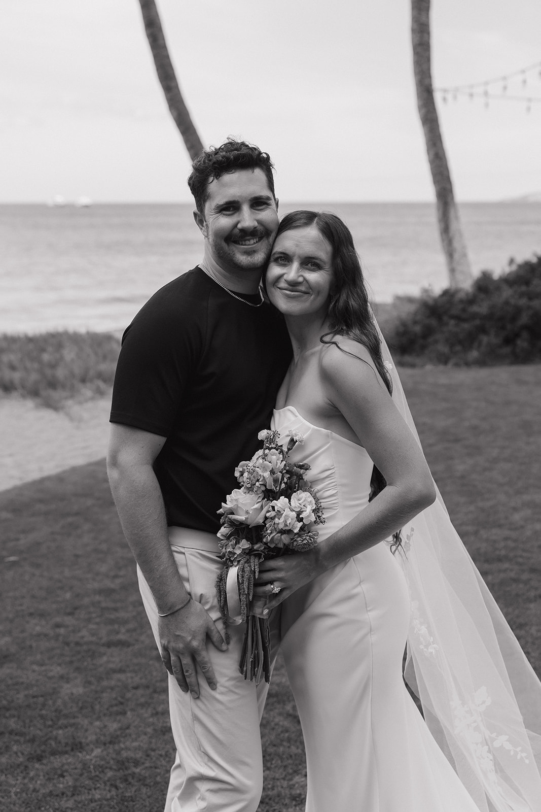Black and white portrait of the bride and groom smiling close together with the ocean and palm trees behind them.