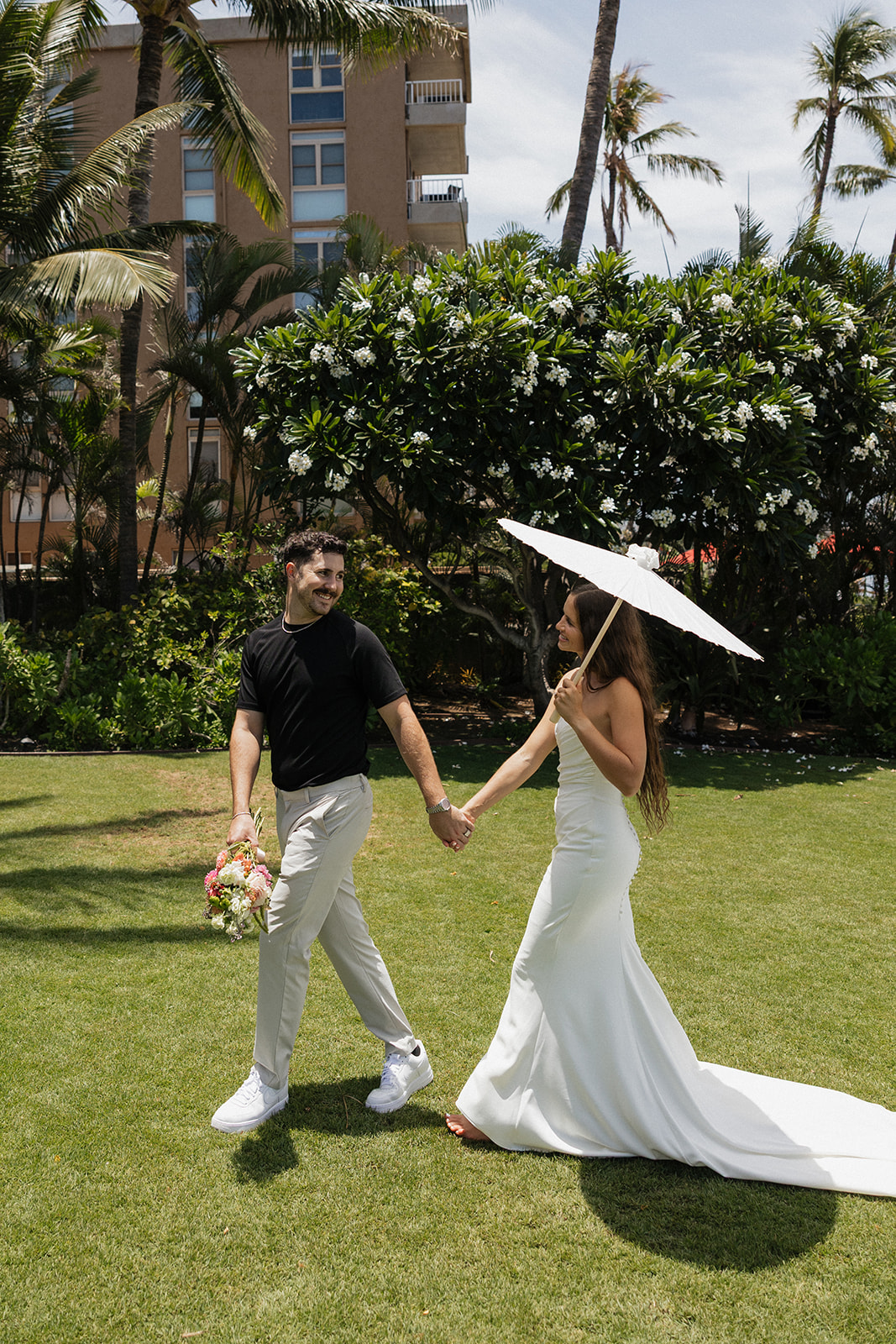 Bride and groom hold hands and smile at each other while walking across the lawn with a white parasol at this Maui wedding venue.