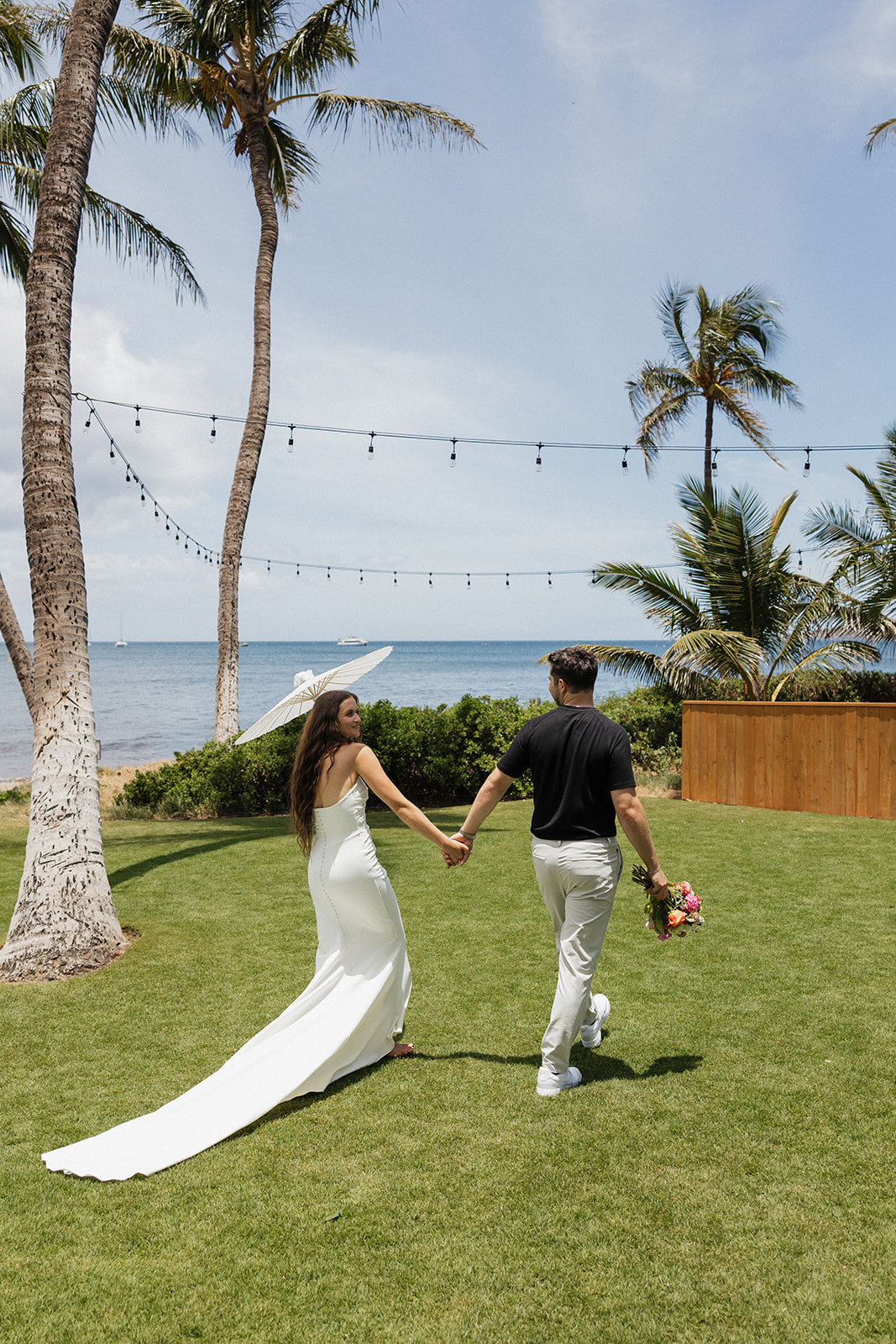 Bride and groom walk hand in hand toward the ocean with a parasol and bouquet at this Maui wedding venue.