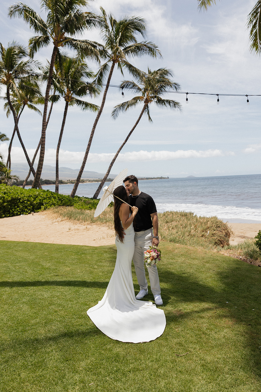 Bride and groom kiss under a parasol on the lawn with palm trees and the ocean glowing softly behind them.