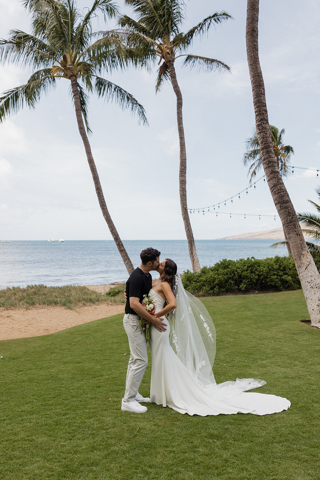 Bride and groom share a kiss under swaying palm trees with the ocean and string lights behind them at this Maui wedding venue.