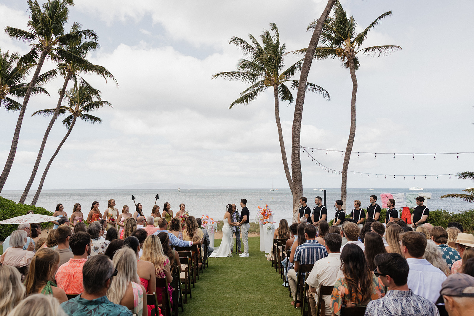 Full ceremony view from behind the guests shows the wedding party, palm trees and ocean at this dreamy Maui wedding venue.