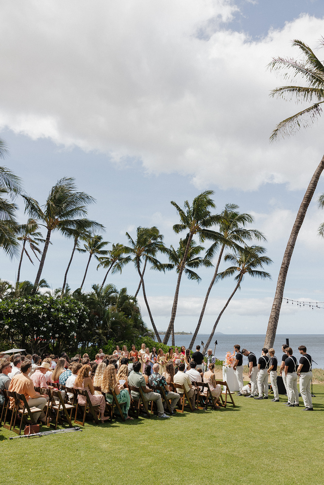 Wide shot of the bridal party, groomsmen and seated guests lined up under palm trees at this oceanfront Maui wedding venue.