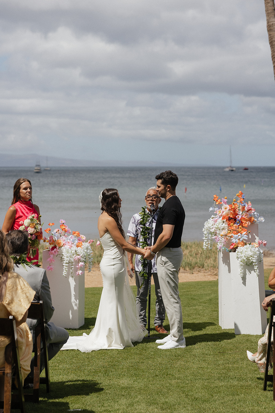 Bride and groom hold hands and share vows with their officiant as the ocean and sailboats stretch out behind them.