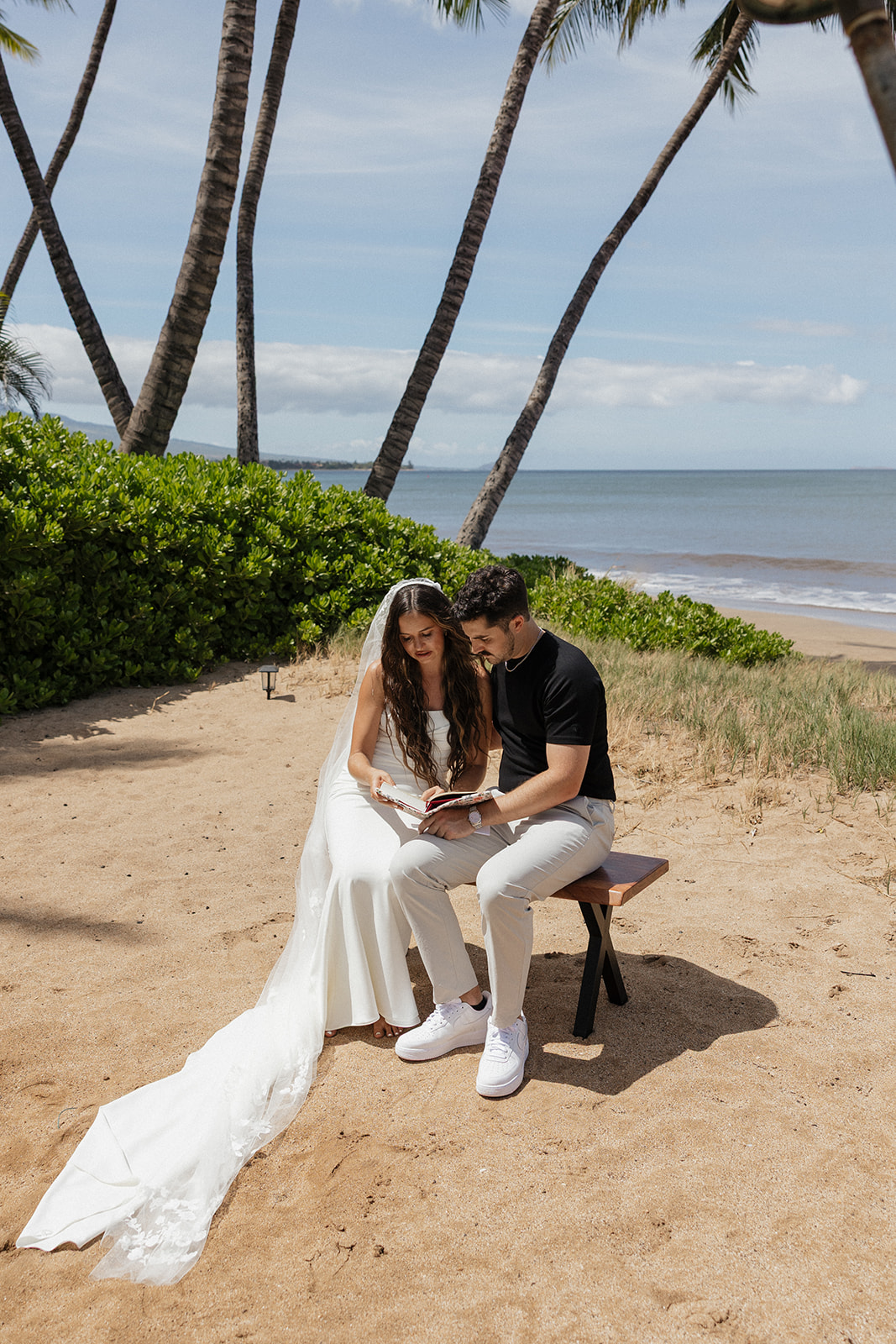 Bride and groom sit on a wooden bench under palm trees reading their vows together at this oceanfront Maui wedding venue.