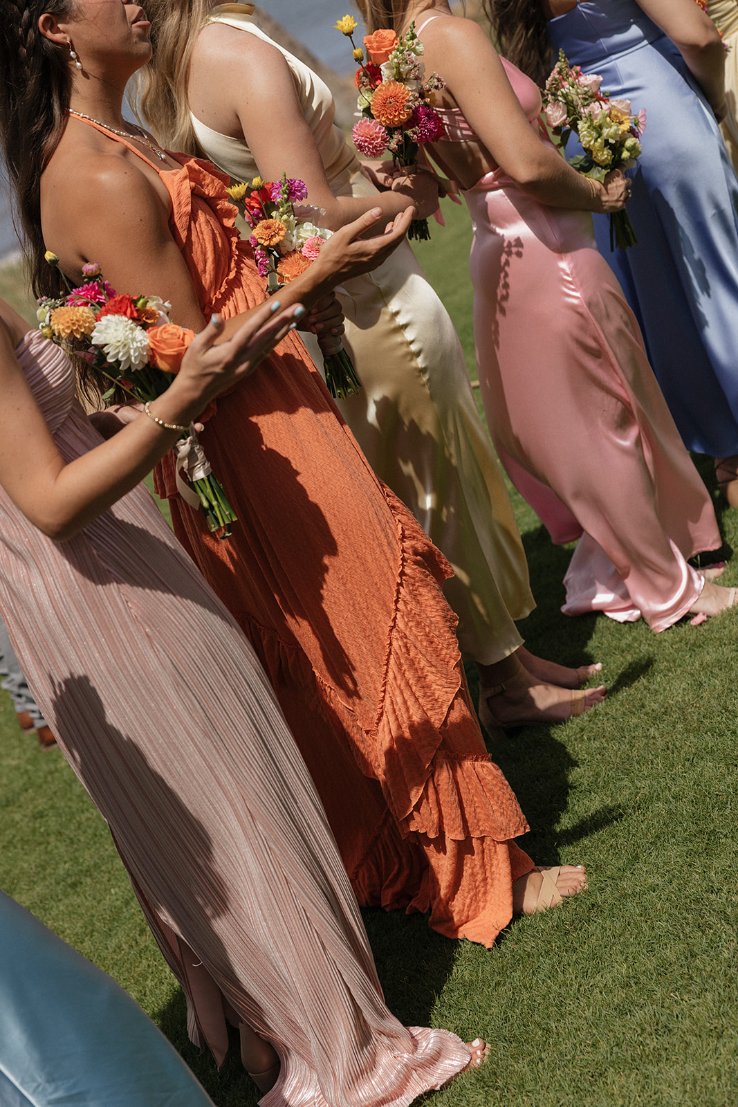 Bridesmaids in orange, pink, yellow and blue dresses hold colorful bouquets during the ceremony at Sugar Beach Events, a stunning Maui wedding venue.