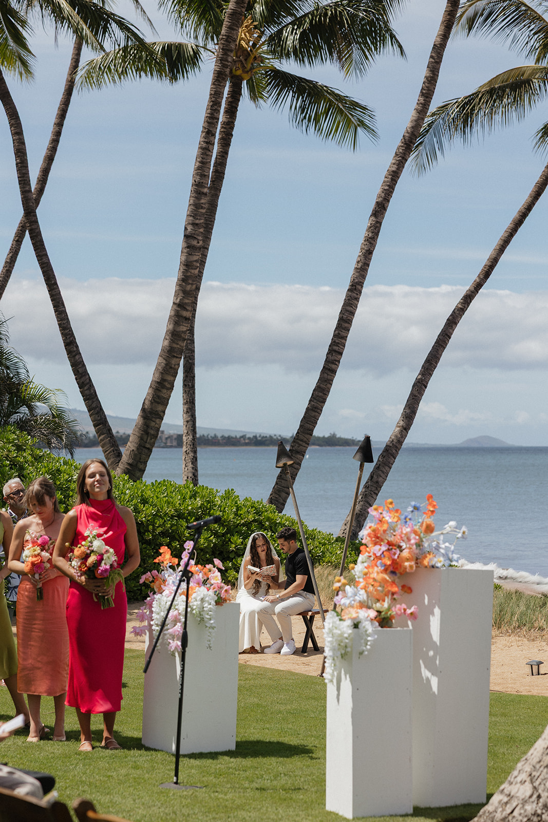 Bridesmaids in pink and orange dresses stand near white floral pillars as the couple shares a quiet moment by the sea.