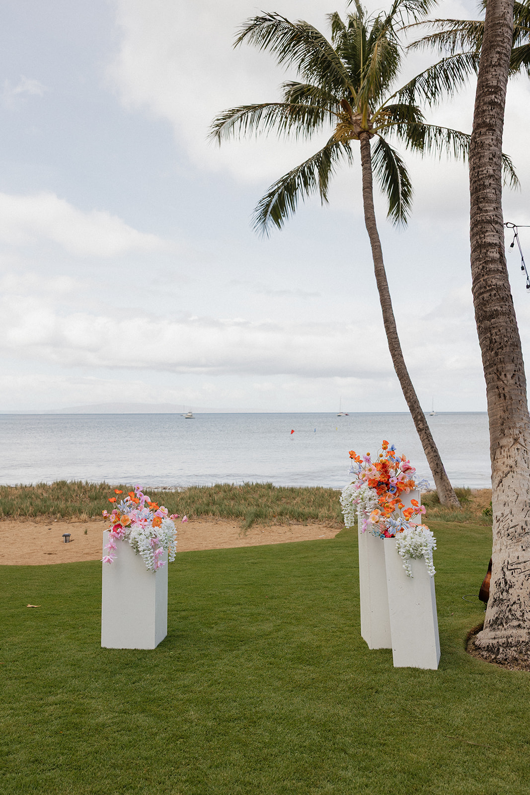 White floral pillar arrangements in orange, blue and pink stand beside a palm tree overlooking the ocean at this Maui wedding venue.