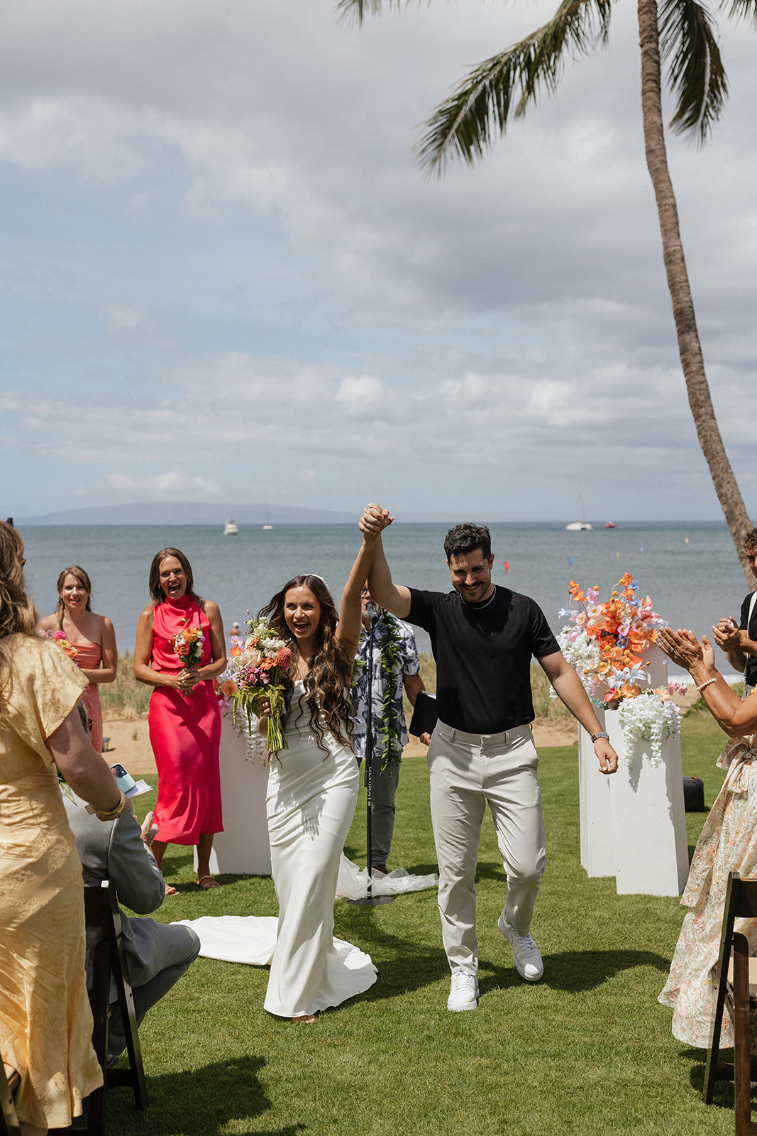 Newlyweds beam and raise their joined hands while walking back down the aisle with the ocean behind them.