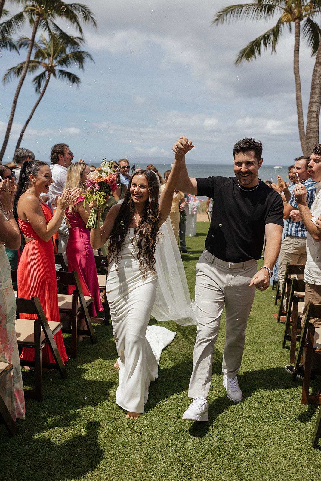 Bride and groom raise their hands and cheer walking back up the aisle through bubbles at this Maui wedding venue.