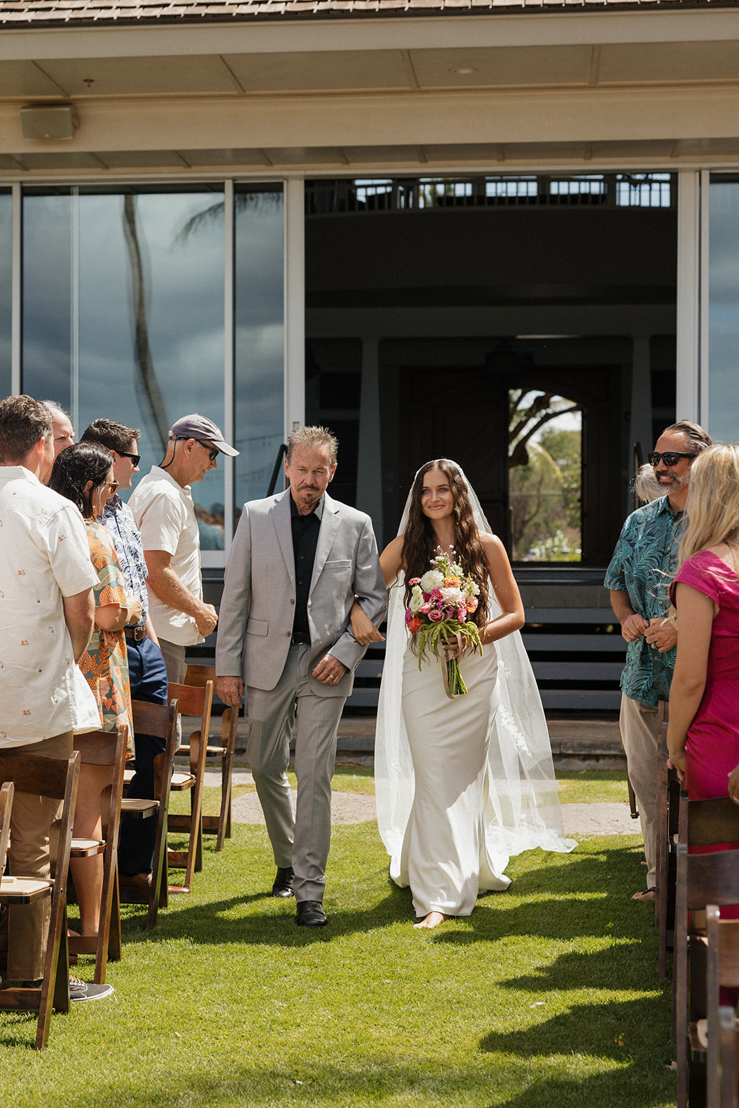 Bride walks down the grass aisle with her father past guests seated in wooden folding chairs at this oceanfront Maui wedding venue.