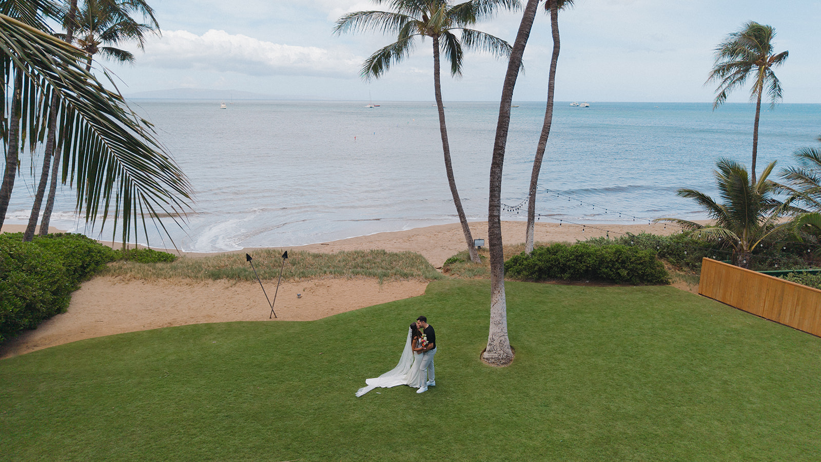 Drone shot captures the bride and groom sharing a kiss on the lawn with the ocean and palm trees framing this Maui wedding venue.