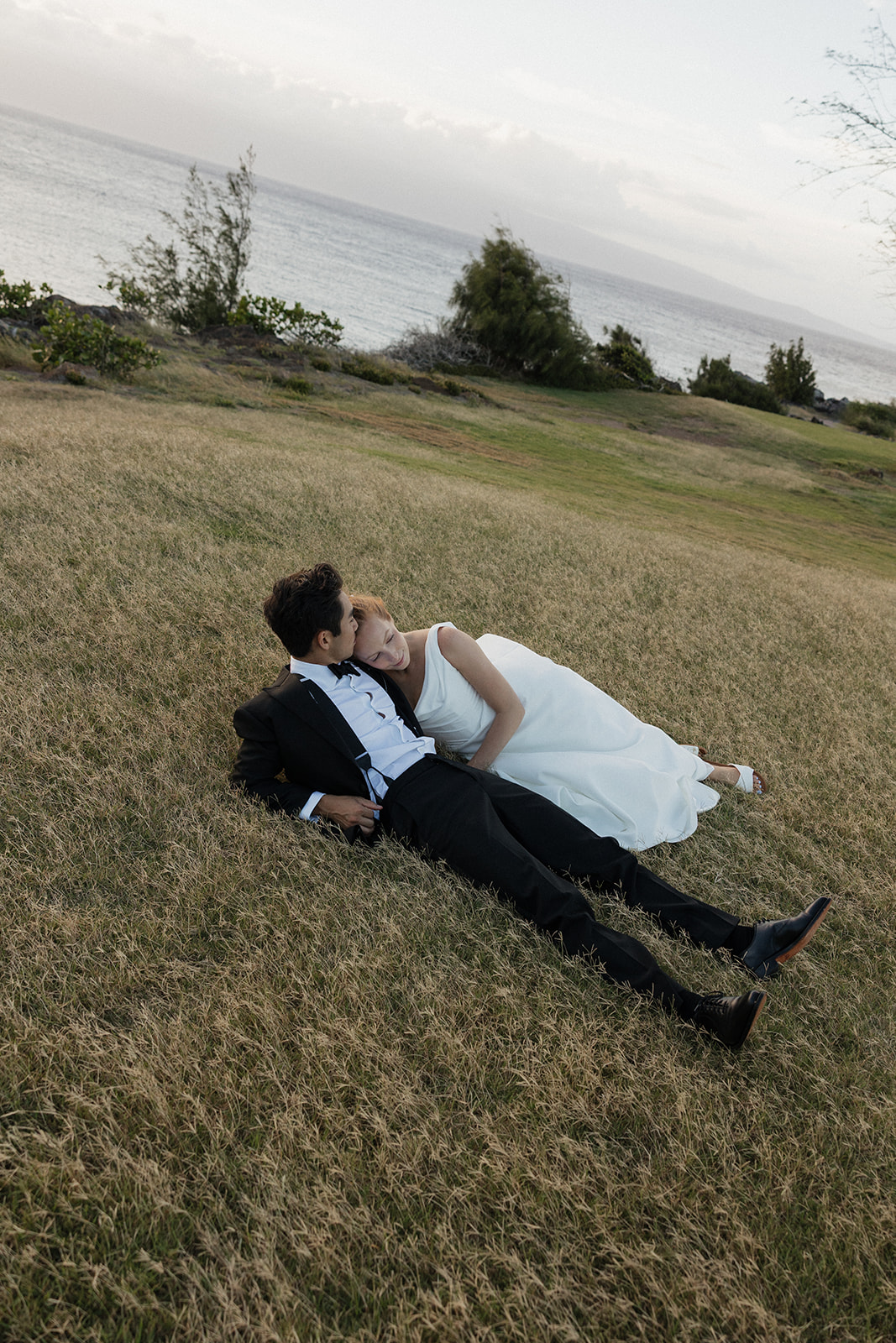 Bride and groom lying together on the grassy hillside above the ocean at sunset near the Maui Pineapple Chapel.