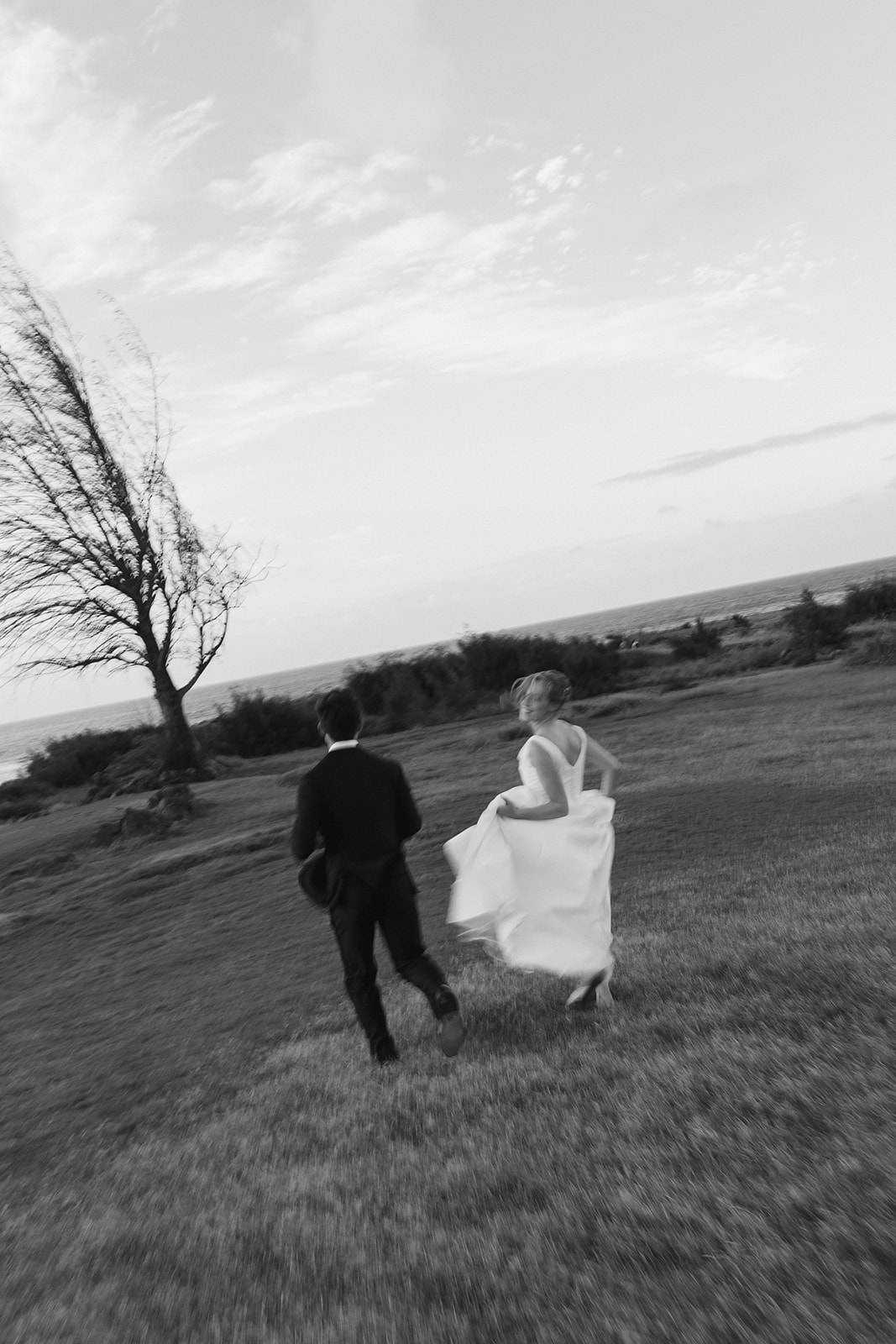 Black and white motion blur photo of a bride and groom running across a windswept field at the Maui Pineapple Chapel.