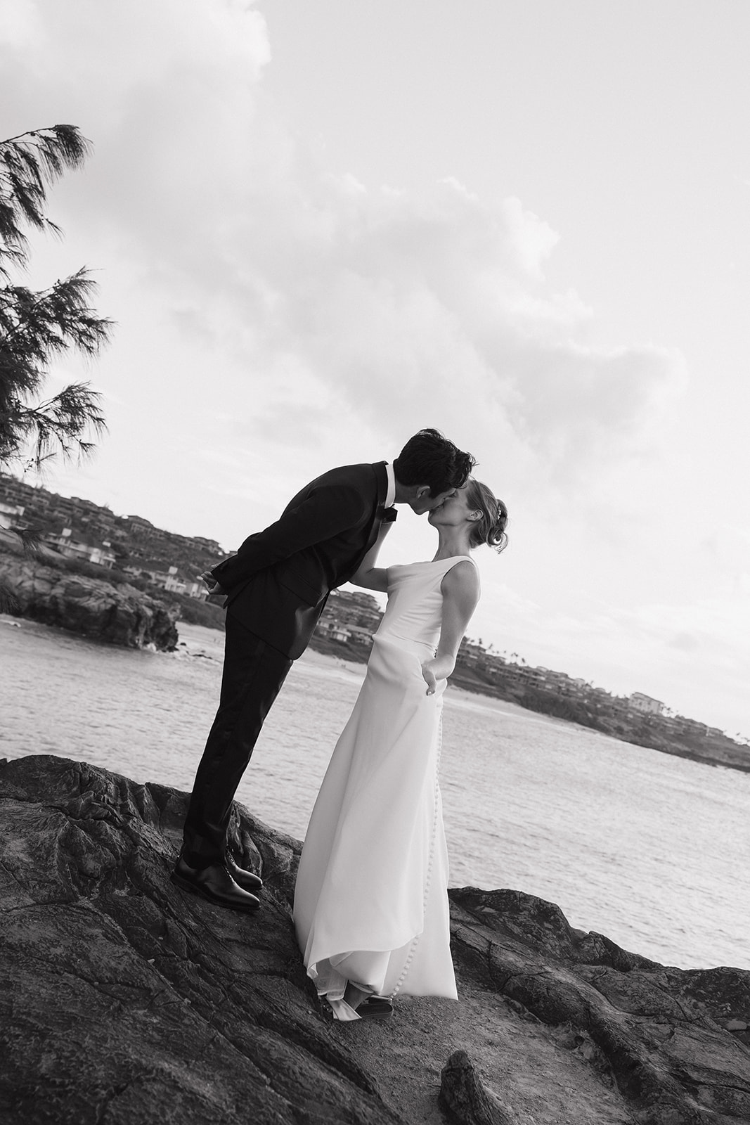 Black and white photo of a bride and groom kissing on coastal rocks above the ocean during their Maui Pineapple Chapel wedding.