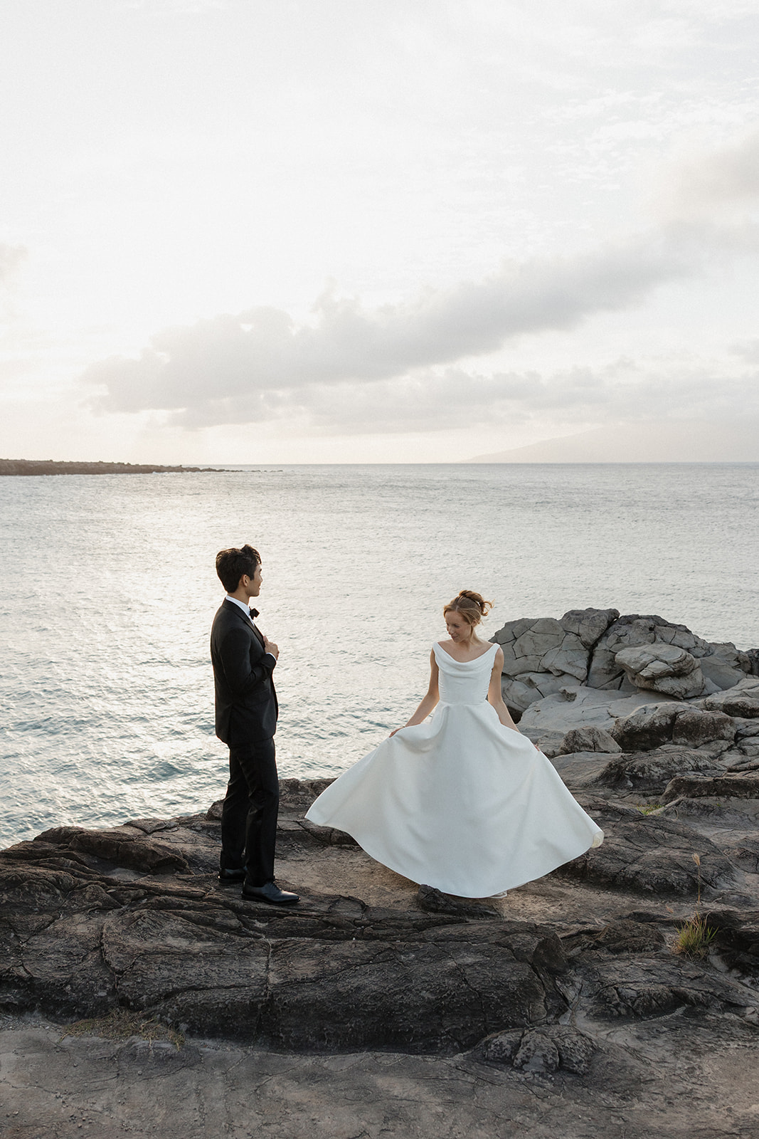 Bride twirling her satin gown on the lava rocks while the groom adjusts his lapel, with the ocean behind them during their Maui Pineapple Chapel wedding.