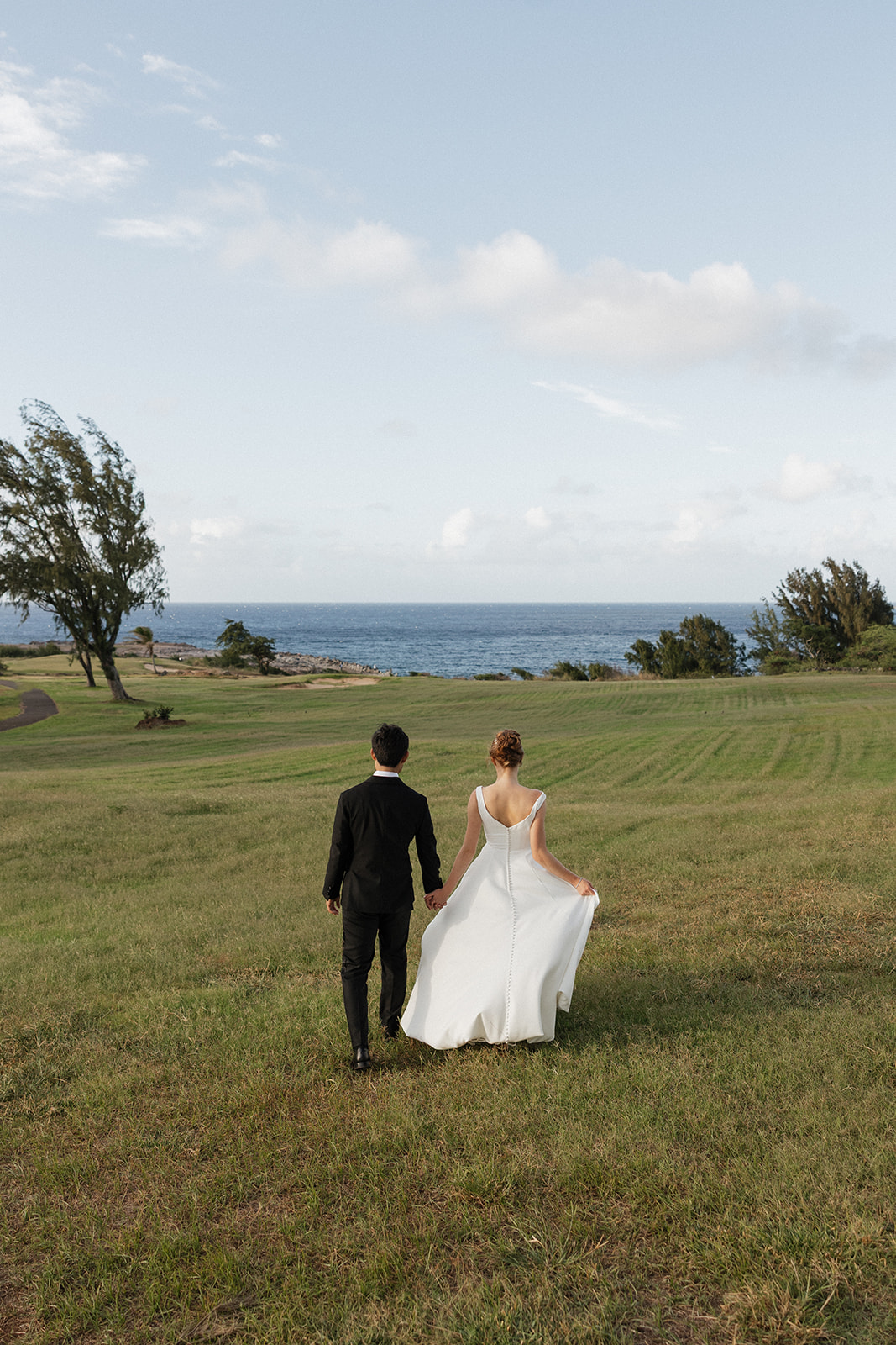 Bride and groom holding hands as they walk across a grassy hillside toward the ocean after their Maui Pineapple Chapel wedding.
