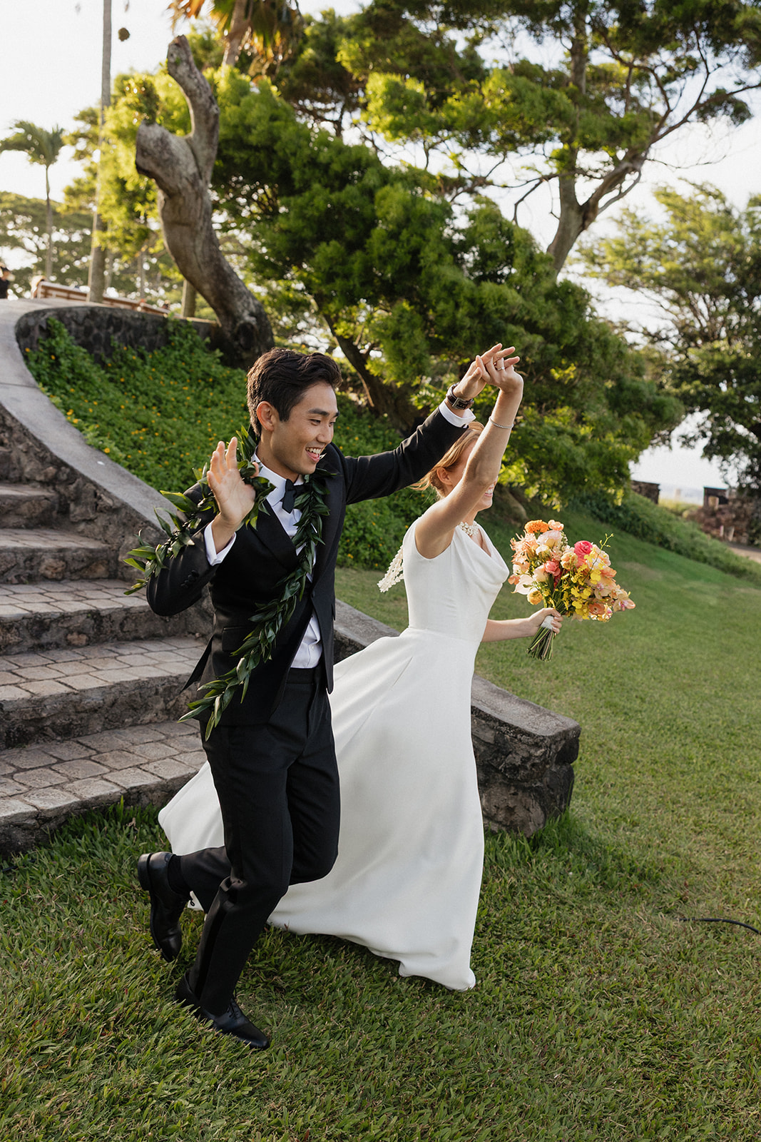 Newlyweds raising their arms and celebrating as they walk down stone steps after their ceremony at the Maui Pineapple Chapel.