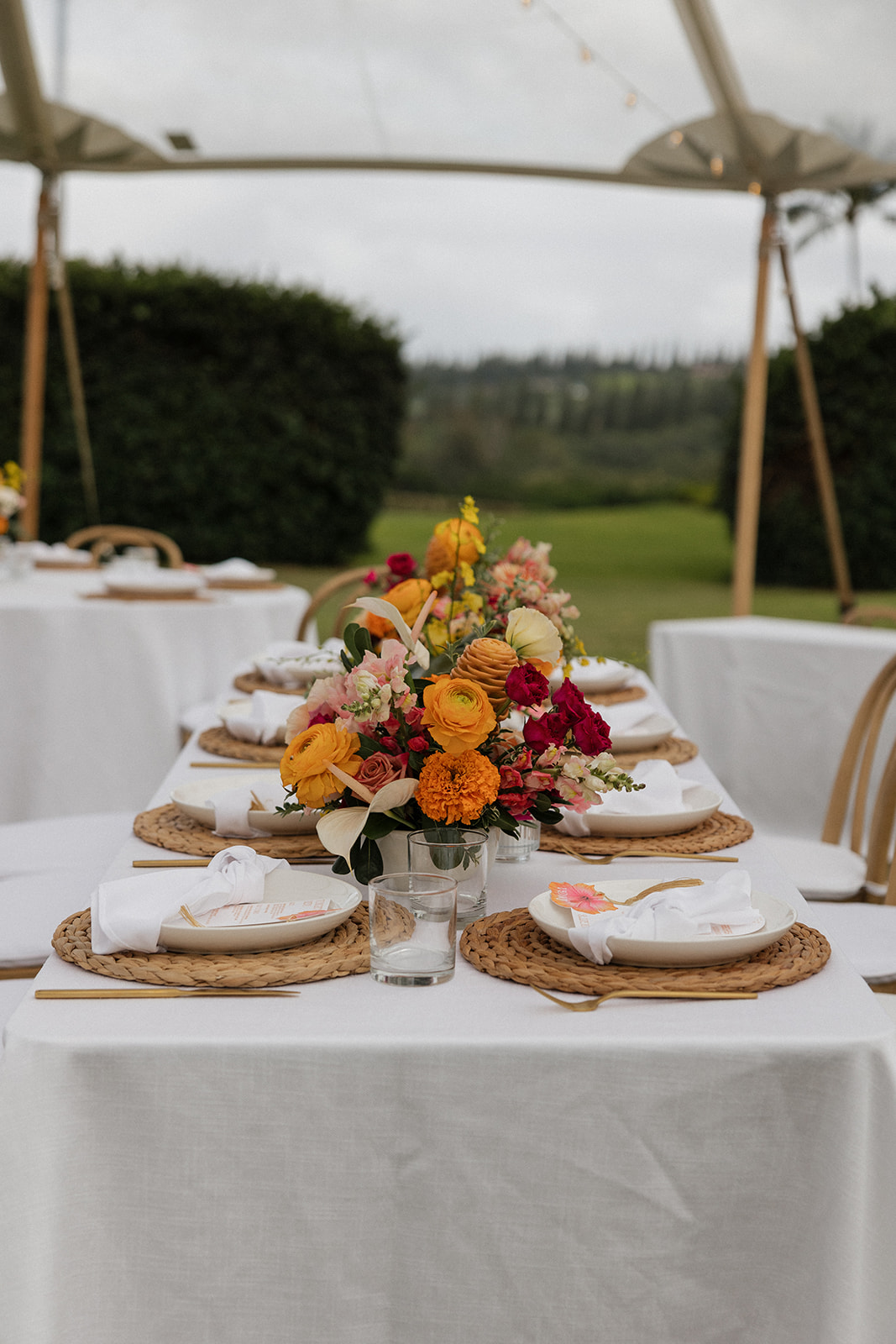 Long reception table with a bright tropical centerpiece, woven placemats, and gold flatware under a tent at the Maui Pineapple Chapel.