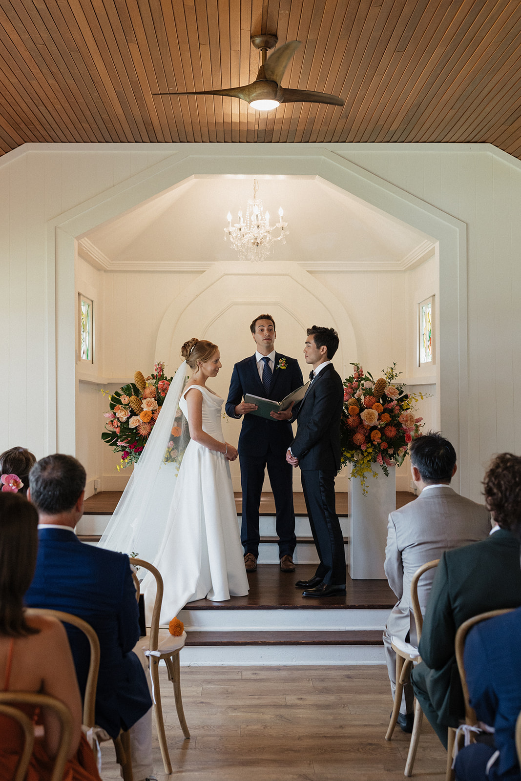 Wide shot of a couple exchanging vows at the altar with tropical floral arrangements and a chandelier inside the Maui Pineapple Chapel.