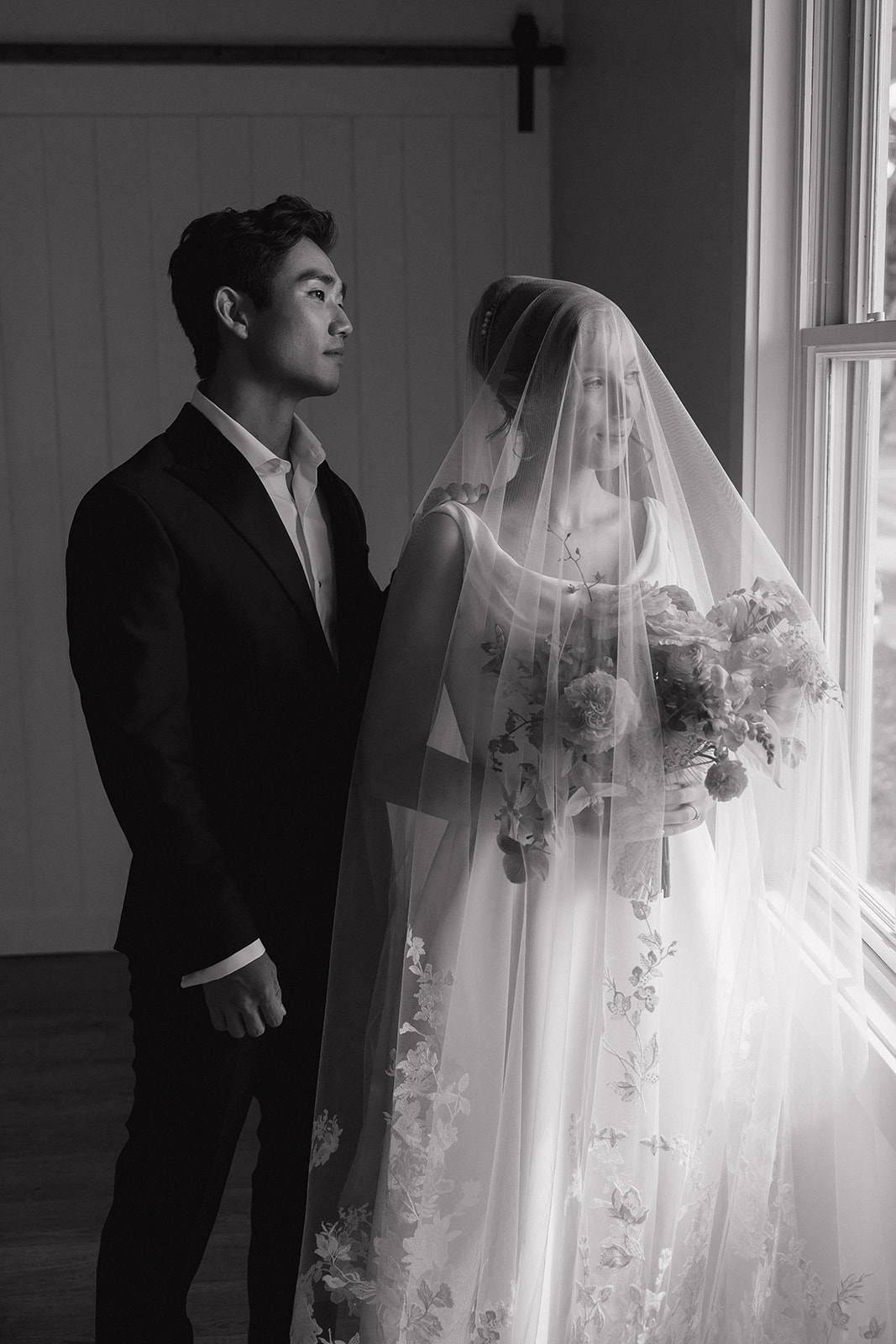 Black and white portrait of a groom standing beside his bride in a lace trimmed veil by the window at the Maui Pineapple Chapel.