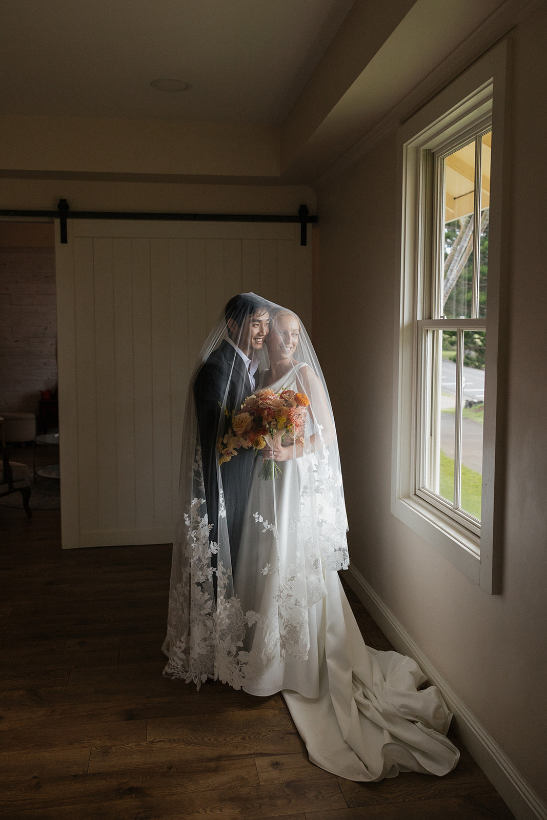 Couple smiling together under a lace veil beside a window and barn door in the bridal suite at the Maui Pineapple Chapel.