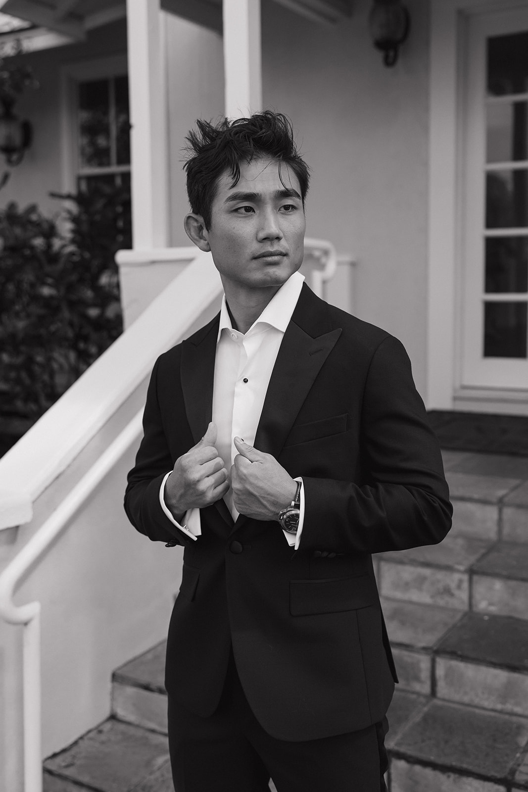Black and white portrait of a groom in a black tuxedo adjusting his lapels on the porch steps of the Maui Pineapple Chapel.
