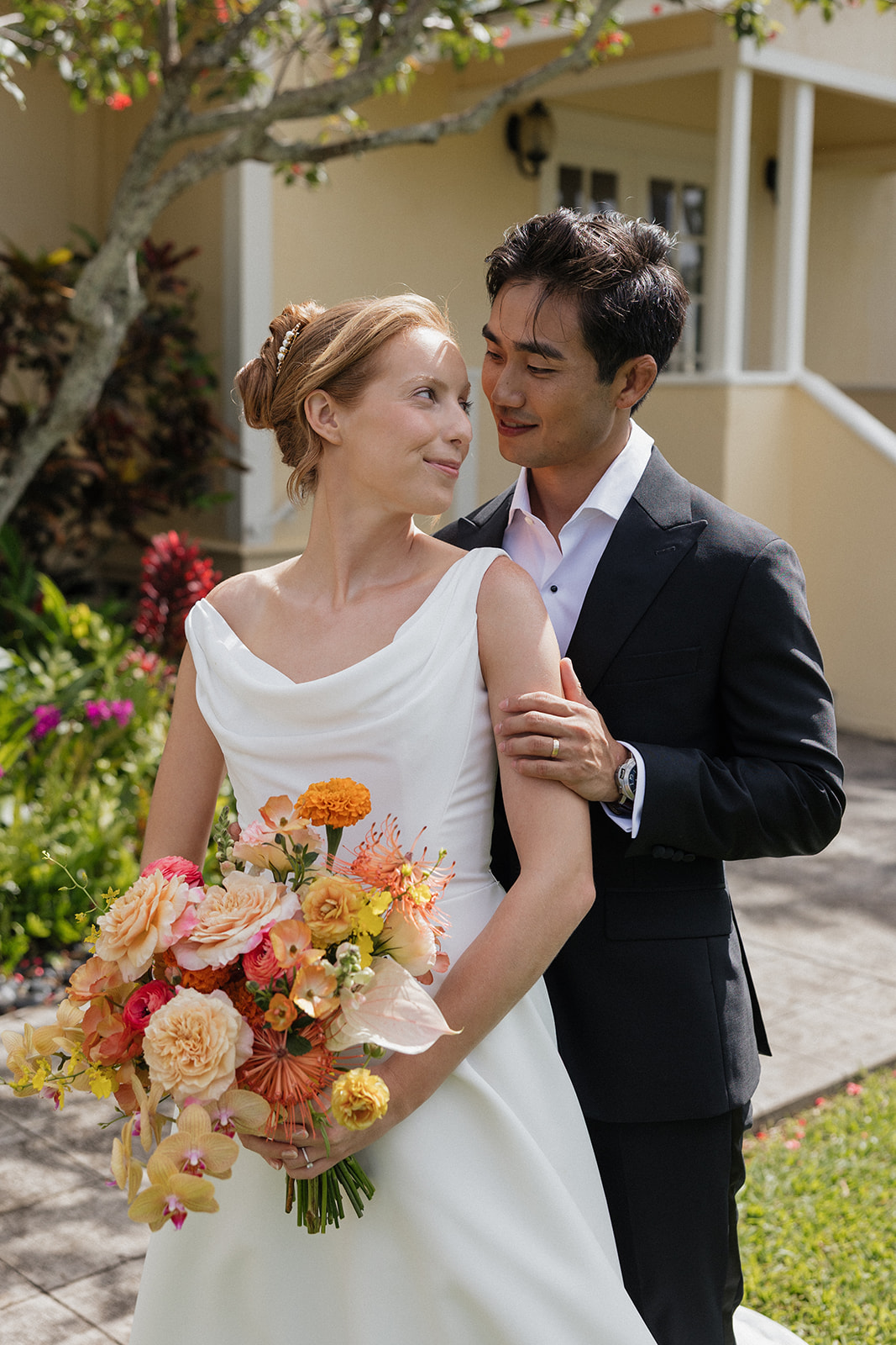 Groom wrapping his arms around his bride as she holds a peach and orange bouquet outside the Maui Pineapple Chapel.