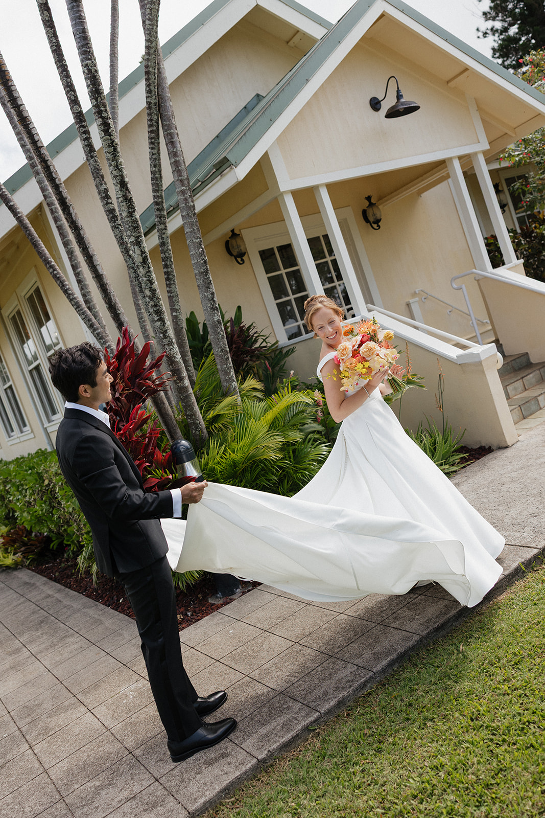 Groom holding the flowing train of his bride's gown while she smiles down at her colorful bouquet in front of the Maui Pineapple Chapel.