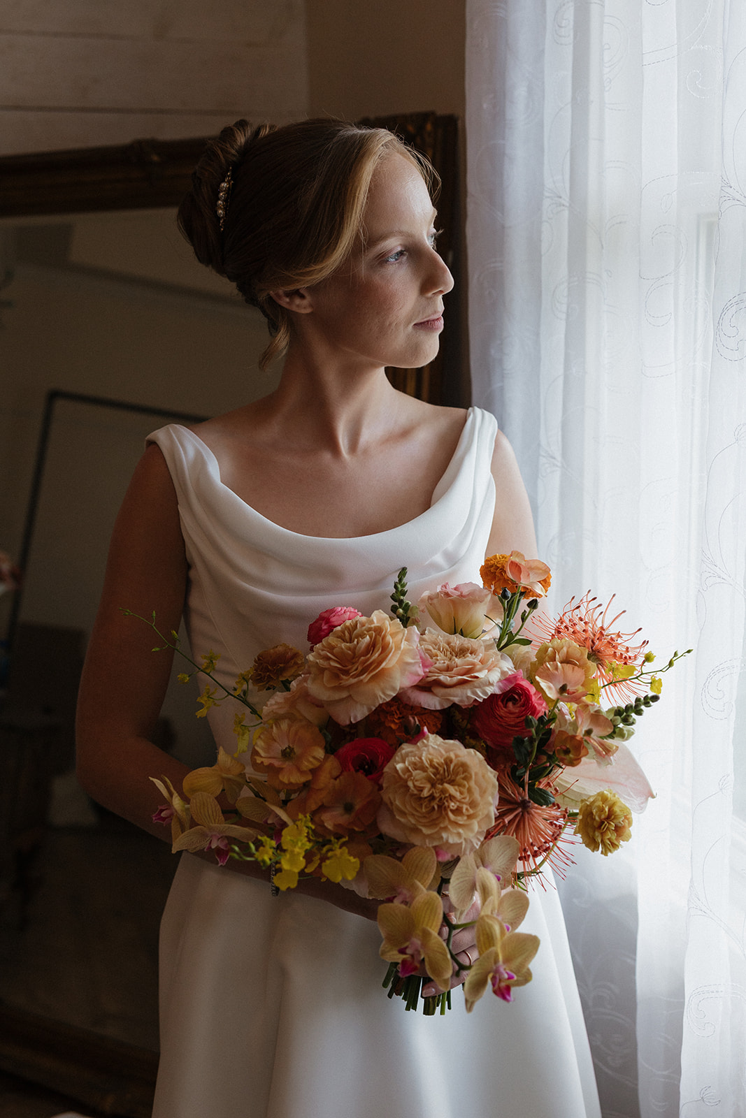 Bridal portrait of a bride holding a peach, coral, and yellow bouquet by a sunlit window at the Maui Pineapple Chapel.