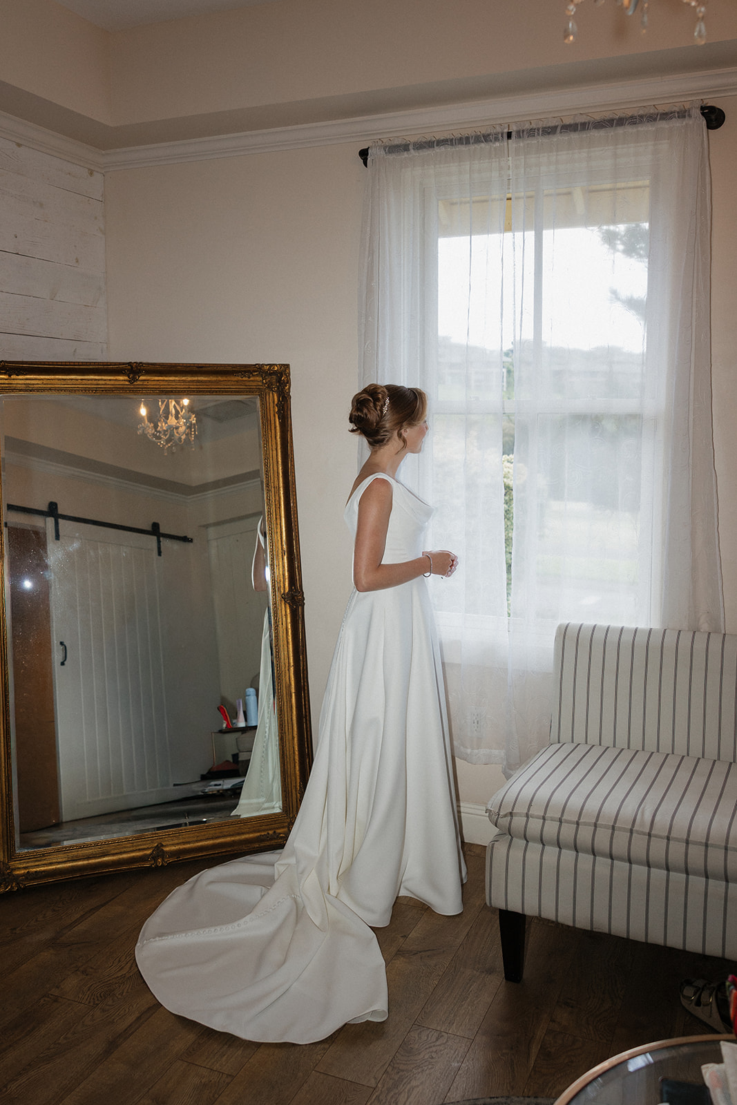 Bride in a cowl neck wedding gown looking out a sheer curtained window beside a gold mirror in the bridal suite at the Maui Pineapple Chapel.