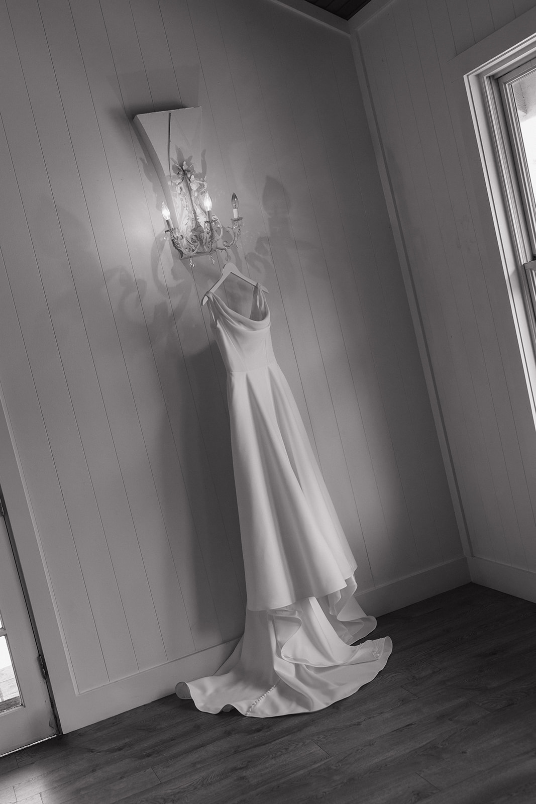 Black and white photo of a satin cowl neck wedding dress hanging from a vintage sconce in the bridal suite at the Maui Pineapple Chapel.