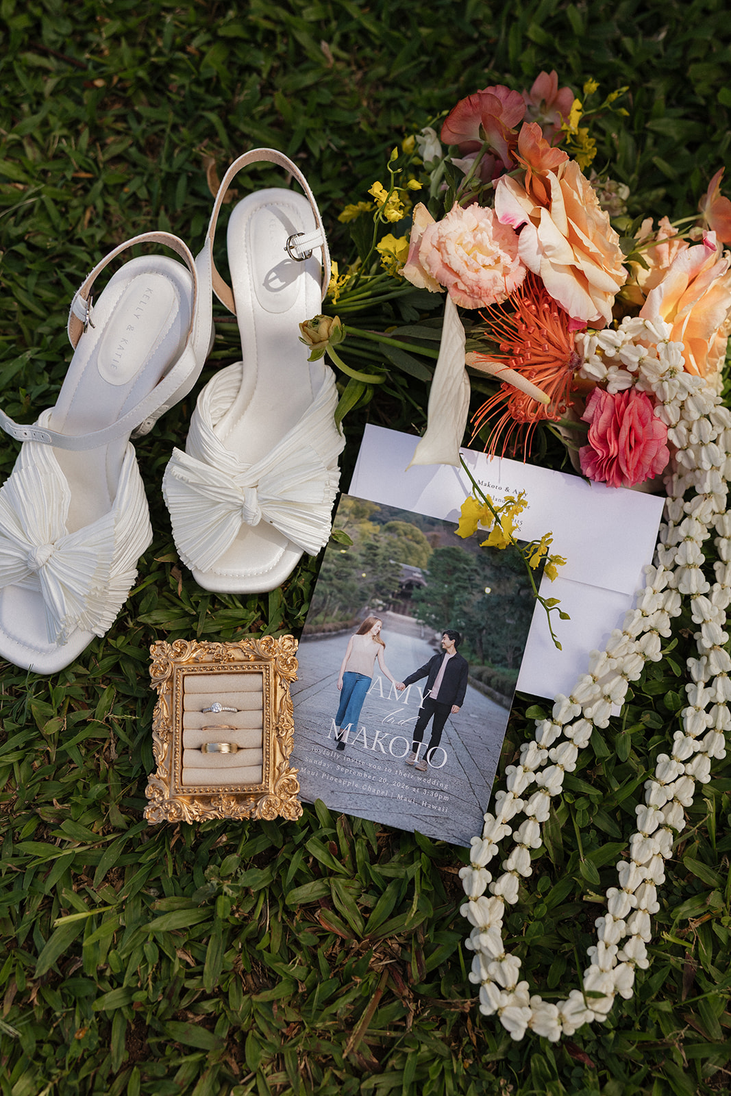 Wedding day details laid out on the grass at the Maui Pineapple Chapel, including white bow heels, rings in a gold frame, an invitation, a tropical bouquet, and a white flower lei.