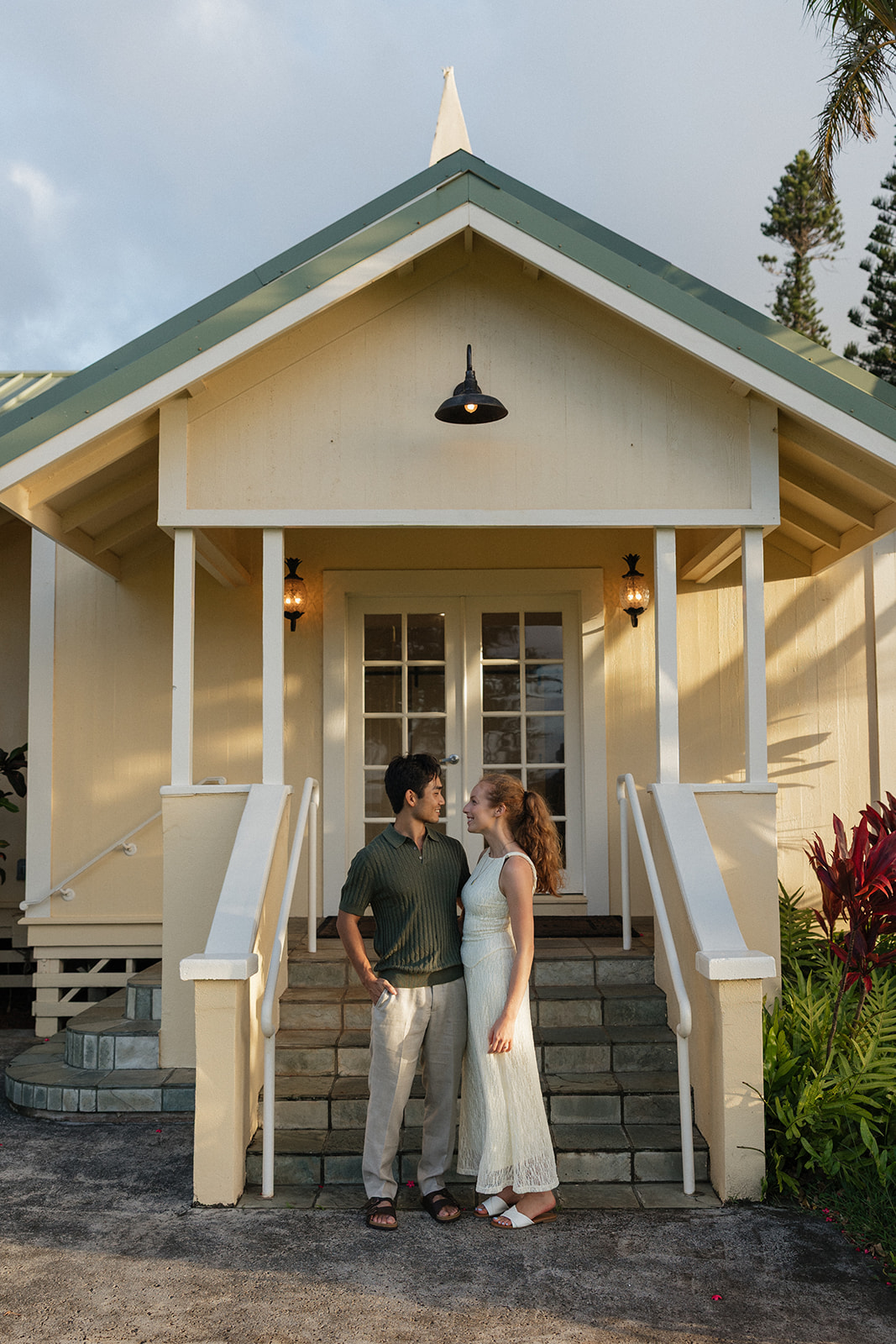 Couple in olive green and cream outfits standing on the steps of the yellow Maui Pineapple Chapel at sunset.